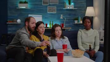 Group of mixed race friends relaxing on sofa while watching entertainment movie on television laughting late at living room during home party. Group of multi-ethnic people enjoying time together video