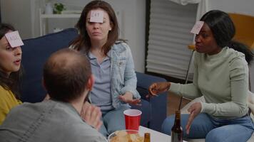 Mixed race friends gathering in living room playing guess who game using sticky notes. Mixed race people having fun, laughing together while sitting on sofa late at night during funny party. video