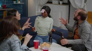 Group of multiethnic friends with sticky notes putting on forehead playing guess who game during home party. Mixed race people having fun, laughing together while sitting on sofa late at night video
