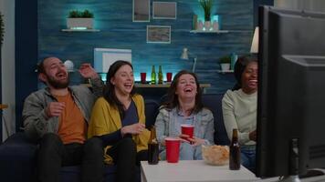 Multiracial friends laughing smiling having fun together while sitting on couch in front of TV watching comedy movie. Group of mixed race people enjoying free time drinking beer eating snacks video