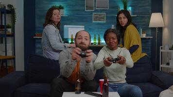 Two women losing online videogames using joystick playing for gaming competition. Multi-ethnic friends drinking beer, socialising, having fun together sitting on couch late at night video