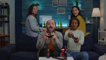 Woman with blak skin playing online videogames against man friends during competition simulation using controller. Group of mixed race winning game while sitting on sofa in living room late at night video