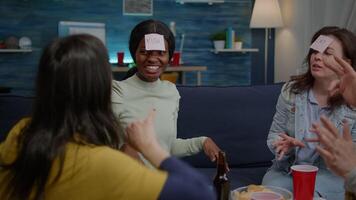 Multi-ethnic friends enjoying time together while relaxing on couch playing guess who game using sticky notes attaching on forehead. Mixed race people having fun in living room late at night video