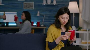 Confident woman reading email notification using smartphone while relaxing on sofa late at night in living room. In background her friends drinking beer, having fun during wekeend party video