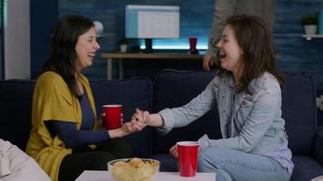 Happy women enjoying friendship with high five late at night in living room during home entertainment party. In background multiethnic friends gathering together having fun drinking beer video