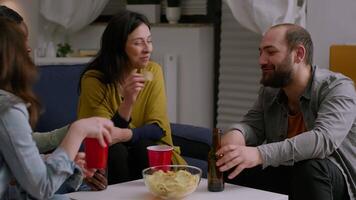 Mixed race friends talking while relaxing on couch in living room late at night drinking beer enjoying home fun party. Group of multi ethnic people hanging out enjoying time spending together video