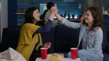 Two women hanging out at home party giving hight five while relaxing on sofa late at night in living room. In background multiethnic friends gathering together having fun drinking beer video