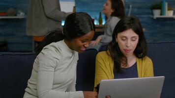 Multi-ethnic friends eating snacks while relaxing on couch watching at comedy on social media using laptop. In background woman and man drinking beer enjoying time spend together video