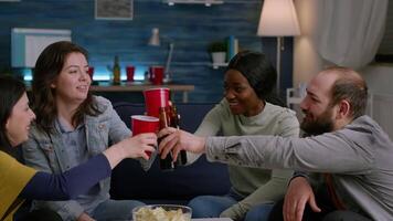 Multi-cultural friends socialising during night fun home party while relaxing on sofa in living room cheering beer bootles. Group of multi-ethnic people hanging out enjoying time spending together video