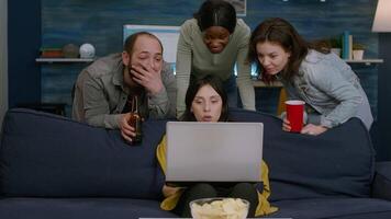 Group of mixed race people spending time together watching comedy movie on laptop computer sitting on sofa late at night in living room. Multiracial friends drinking beer, talking during relax wekeend video