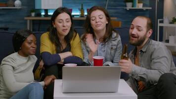 Happy multi-ethnic friends greeting her collegue during online videocall meeting using laptop webcam. Group of multiracial people spending time together sitting on couch late at night in living room video