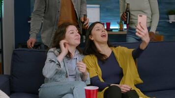 Group of mixed race people taking pictures with phone while sitting on couch in living room spending time together. Multiracial friends posting selfie on internet sharing with other person video