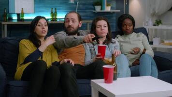 Multi-ethnic friends relaxing on sofa late at night during home party changing channel on television using remote while drinking beer. Group of multiracial people enjoying time together video