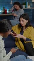 Vertical . Mixed race friends sitting on sofa holdings mobile in hands browsing on internet, watching online during relaxing party. Group of multi-ethnic hanging out together spending time in living room video