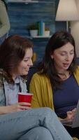 Vertical . Multi-ethnic women enjoying time together watching at movie on laptop while resting on couch. In background woman and man drinking beer, laughing late at night during relaxing party video