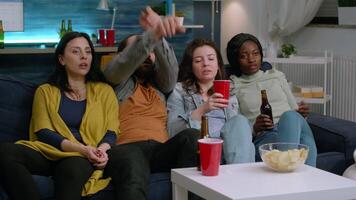 Multi-cultural friends resting on couch late at night during home party watching entertainment movie on television drinking beer. Group of multiracial people enjoying time together video