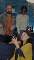 Vertical . Multiracial friends hanging out together socializing while looking at funny on smartphone. Group of multi-ethnic people sitting on sofa late at night in living room drinking beer video