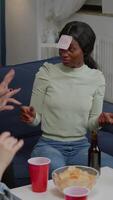 Vertical . Group of multiethnic friends with sticky notes putting on forehead playing guess who game during home party. Mixed race people having fun, laughing together while sitting on sofa late at night video
