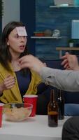 Vertical . Mixed race friends playing Guess who game with sticky papers attached on forehead. Group of multiracial people having fun, laughing together while sitting on sofa in living room late at night video