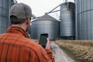 A farmer attentively checks a smartphone while overseeing the grain storage silos on the farm, highlighting the significant intersection of agriculture and technology in modern farming practices photo