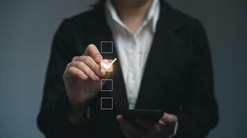 A woman in a business suit is holding a cell phone and drawing a checkmark on virtual checkbox. Concept of professionalism and accomplishment, quality check list business concept. photo