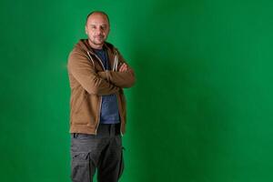 Middle aged bald man in casual attire with crossed arms poses confidently against a green studio backdrop. photo