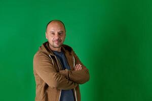 Middle aged bald man in casual attire with crossed arms poses confidently against a green studio backdrop. photo