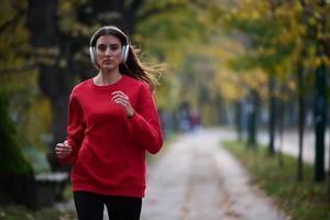 Young beautiful woman running in autumn park and listening to music with headphones on smartphone photo