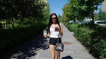 Woman walking in a park holding a drink while using her smartphone on a sunny day video