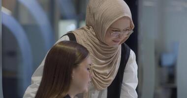 A diverse group of businesswomen, one of whom wears a hijab, collaborates while focusing on a laptop, engaging in discussions and problem-solving, showcasing professionalism, teamwork, and effective video