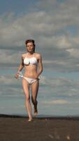 lleno longitud ver de mujer atleta corriendo en arenoso playa. esbelto mujer vestido en blanco bikini traje de baño durante verano playa formación en soleado día. frente vista, Mano disparo, vertical real hora video