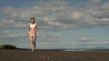 Slim woman athlete running on sandy beach. Sensuality woman dressed in white bikini swimsuit during summer beach training in sunny weather. Front view, handheld shot, real time video