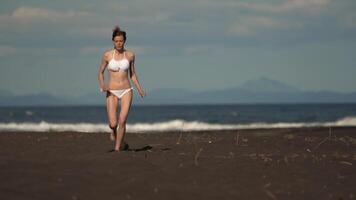 Inspirational slender fitness model running on sandy beach. Sportsperson wearing white bikini swimwear during summer beach training on sunny day. Front view, handheld shot, real time , zoom out video