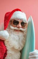 Santa Claus wearing sunglasses, holding a surfboard, smiling on a bright pink background photo