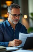 Middle aged man sitting at his desk, reading documents while using a laptop, looking focused photo