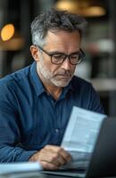 Middle aged man sitting at his desk, reading documents while using a laptop, looking focused photo