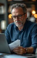 Middle aged man sitting at his desk, reading documents while using a laptop, looking focused photo