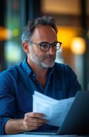Middle aged man sitting at his desk, reading documents while using a laptop, looking focused photo