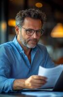 Middle aged man sitting at his desk, reading documents while using a laptop, looking focused photo