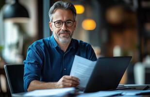 Middle aged man sitting at his desk, reading documents while using a laptop, looking focused photo