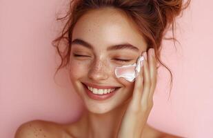 A lovely woman with closed eyes smiles while applying cream to her cheek with one hand photo