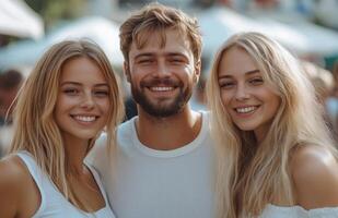 Young man smiling, standing between two blonde women in white outfits on an urban street photo
