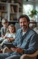 Happy father with a tablet, sitting on a chair in a modern living room, children playing nearby photo