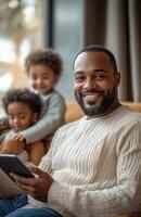 Happy father with a tablet, sitting on a chair in a modern living room, children playing nearby photo