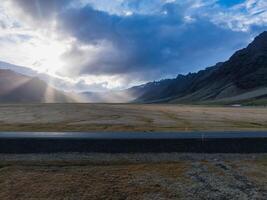 A paved road cuts through a vast, flat plain in Iceland, with mountains in the distance. The cloudy sky and sunlight create dramatic lighting and long shadows. photo