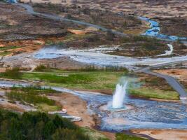 Stunning aerial shot of a geyser erupting in Iceland, surrounded by a pool of water and steam, with a winding river and path leading to it. photo