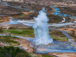 An aerial shot capturing a geyser erupting in Iceland, surrounded by a muddy area and a small stream. A winding road and a small forest are also visible. photo