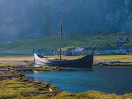 A wooden longboat with a high prow and stern sits in calm blue waters. The rocky hillside, covered in green grass and dotted with small buildings, completes the serene Icelandic scene. photo