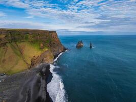 Stunning aerial view of a black sand beach in Iceland, bordered by a steep cliff and deep blue ocean. White waves crash on the shore with rock formations in the distance. photo