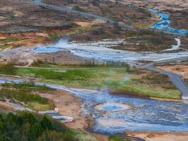 A stunning aerial view of two vibrant blue geysers surrounded by a barren landscape with a winding river in Iceland. The mix of brown, green, and blue creates a striking contrast. photo